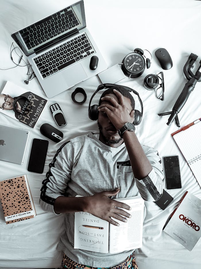 our-services-2 A young man lying on a bed surrounded by modern gadgets, books, and tech devices, illustrating digital lifestyle.
