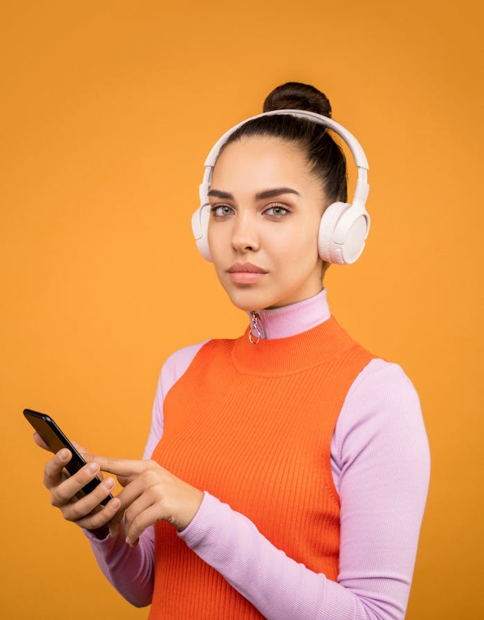 why-choose-us A young woman listens to music on wireless headphones while using a smartphone against an orange background.
