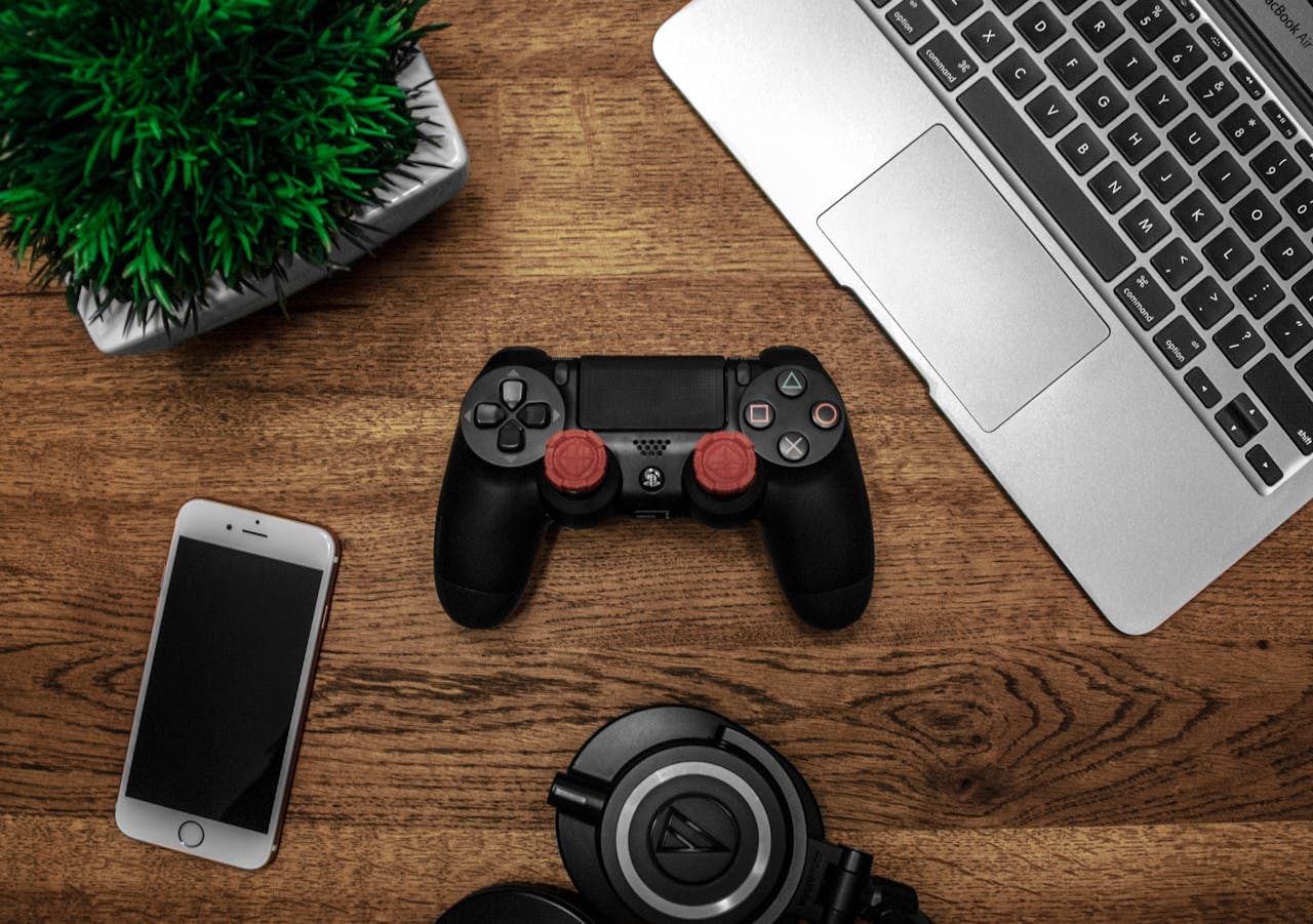 hero-img-01 A flat lay photo showcasing a gaming console, laptop, smartphone, and headphones on a wooden tabletop.