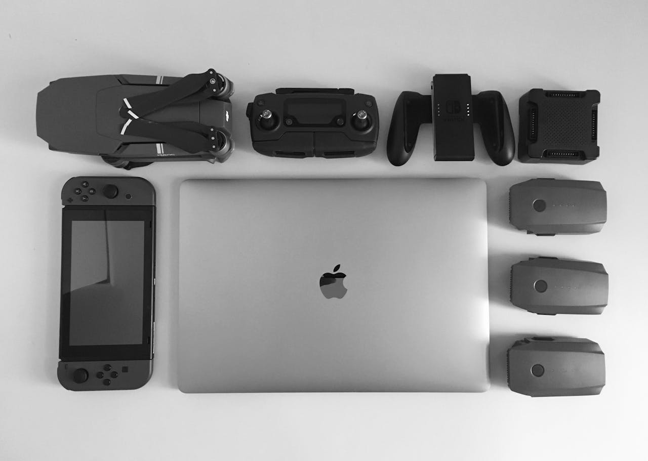 hero-img-02 A black and white flat lay of various modern tech gadgets including a laptop, drone, and gaming device.
